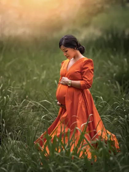 A solo outdoor portrait of an expecting mother in a vibrant orange dress, standing amidst tall green grass. The soft, golden light gives the image a dreamy, ethereal quality.