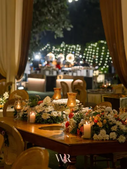 A close-up of a dining table at the rustic-themed event. The warm glow of candles in mercury glass holders complements the wooden table and colorful floral arrangements.