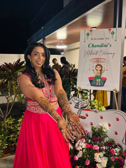 The bride posing next to her mehendi ceremony welcome sign, proudly showing off her intricate henna art.