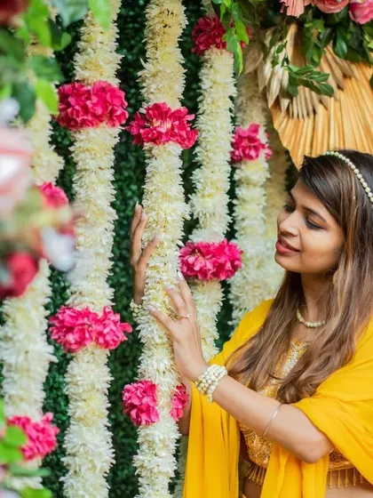 A bride enjoying the beautiful floral backdrop at her Haldi. The contrast between the green wall, white and pink flowers, and her yellow outfit is simply stunning.