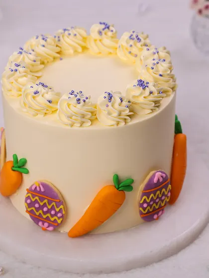 We decorate for the holiday with this festive Easter cake. A smooth, cream-colored cake is adorned with colorful, hand-decorated Easter egg cookies and fondant carrots, creating a bright and cheerful design.