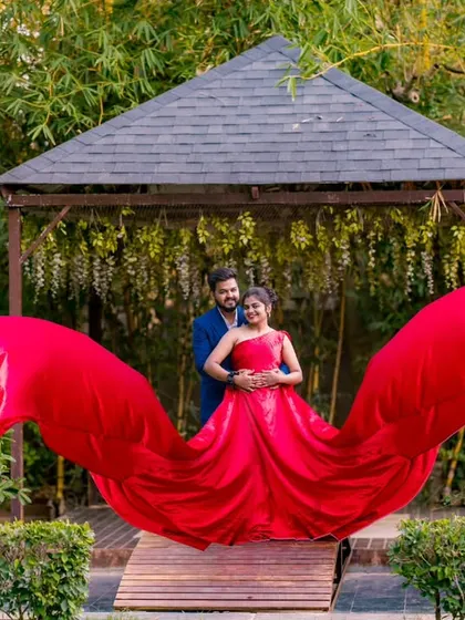 The vibrant red gown with its wing-like sleeves creates a stunning visual against the natural backdrop of the gazebo. This is a perfect example of a high-fashion couple portrait.