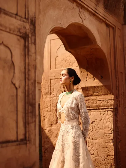A model styled in an ethereal ivory lehenga with delicate cutouts. The natural light and historic setting enhance the romantic and sophisticated feel of this look, designed for a modern bride.