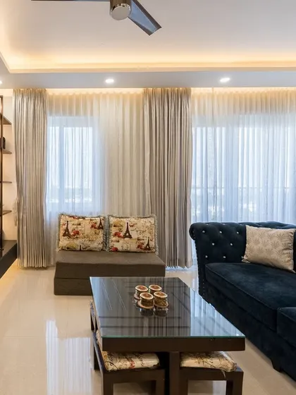 A comfortable living room with a dark blue tufted sofa and a floating TV unit. The sheer curtains allow for ample natural light, creating a bright and welcoming atmosphere.