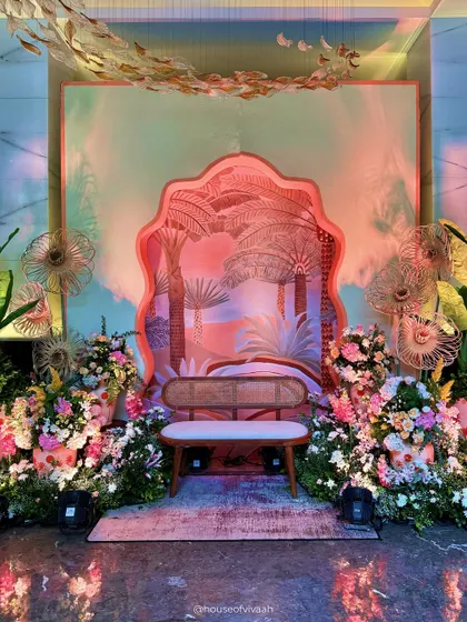 A whimsical photo backdrop for the Welcome Lunch, featuring a layered, scalloped arch with a palm tree motif, surrounded by lush florals and artistic fan-like structures.