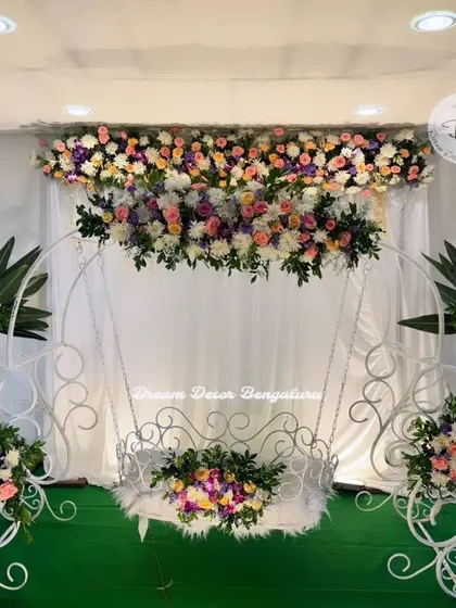 An elegant cradle ceremony setup with a white metal swing. The stage is beautifully decorated with pastel floral arrangements on stands, creating a clean and sophisticated look.
