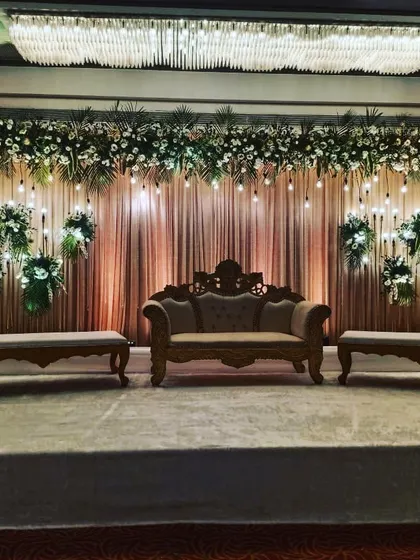 An elegant reception stage with a draped backdrop and hanging Edison bulbs. The floral arrangements and classic carved sofa add a touch of rustic charm and sophistication to the decor.