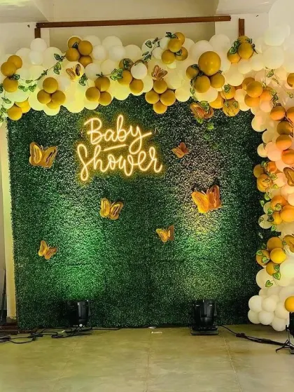 A lush green hedge wall serves as the perfect backdrop for this baby shower celebration. I used a warm neon sign, a flowing organic balloon garland in yellow and white, and delicate butterfly accents to create a cheerful and nature-inspired theme.