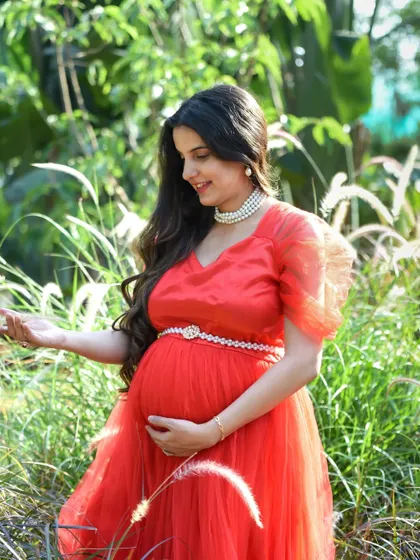 A soft and dreamy outdoor portrait. The mom-to-be, in a lovely red gown, is standing in a field of tall, feathery grass, creating a very gentle and natural look.