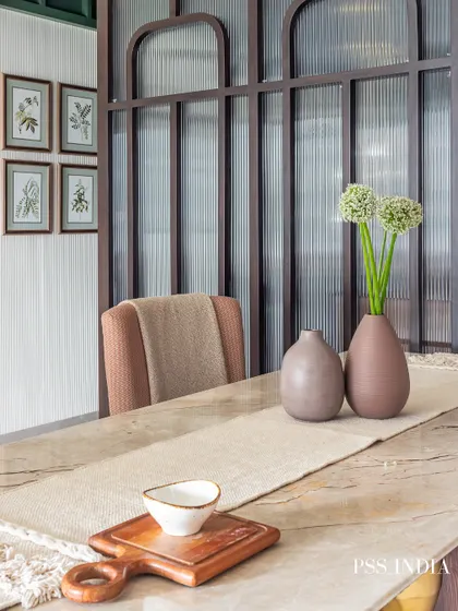 A close-up of a beautifully styled dining table. A simple linen runner, minimalist ceramic vases, and a wooden serving board create an earthy and inviting tablescape, complemented by fluted glass partitions in the background.