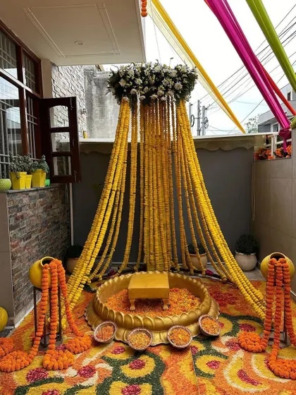 An intimate Haldi setup perfect for a home balcony or small space. It features a canopy of marigold strings and a floral rangoli on the floor, making the occasion special even at home.