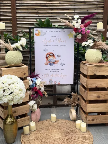 A personalized welcome sign for a baby shower, set against a rustic arrangement of wooden crates, candles, and floral arrangements with pampas grass.