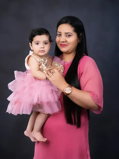 A mother holds her daughter, both looking at the camera. The mother's pink dress and the baby's sparkly outfit make for a sweet and stylish portrait.