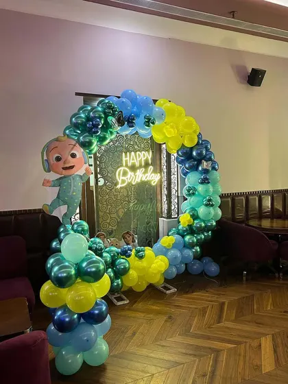 A Cocomelon-themed birthday setup in a restaurant or lounge. A circular balloon arch in shades of green and blue frames a doorway.