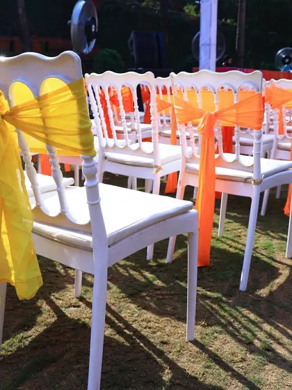 Simple and bright guest seating. White chairs are decorated with yellow and orange organza bows, adding a splash of color that ties in with the vibrant Haldi theme.