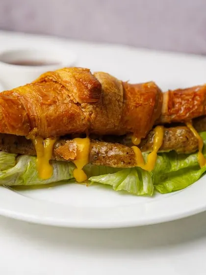 A close-up of a sausage croissant sandwich, highlighting the texture of the bread and the melted cheese. This clean shot is designed to make the item look as tasty as possible for online ordering.