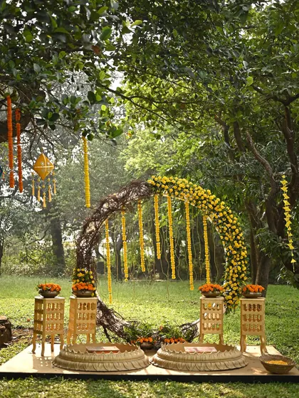 A rustic and natural Haldi setup in a wooded area. The centerpiece is a circular arch made of twigs and decorated with marigold garlands, creating a beautiful blend of rustic charm and traditional elements.