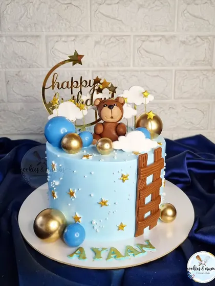 Teddy in the clouds. This dreamy blue cake is decorated with a fondant teddy, stars, clouds, and a ladder, creating a whimsical scene for a little one's birthday.