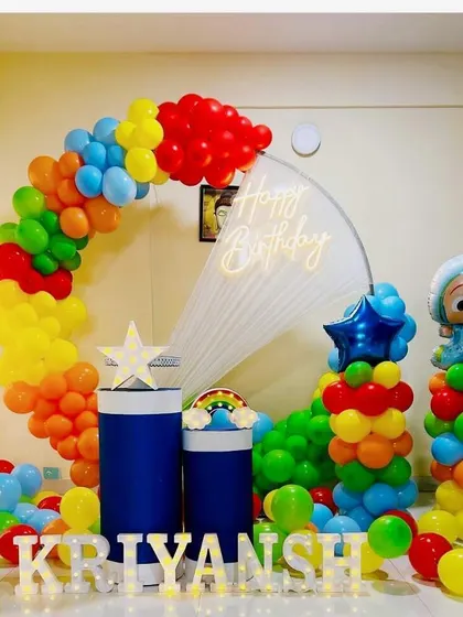 A rainbow-colored Cocomelon birthday setup. The circular backdrop is framed by a rainbow balloon garland, with character props and light-up marquee letters in the foreground.