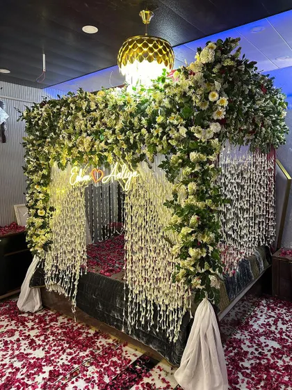 An opulent wedding night decoration with a bed completely enveloped in a canopy of white flowers and hanging floral strings. The floor is covered in red rose petals.