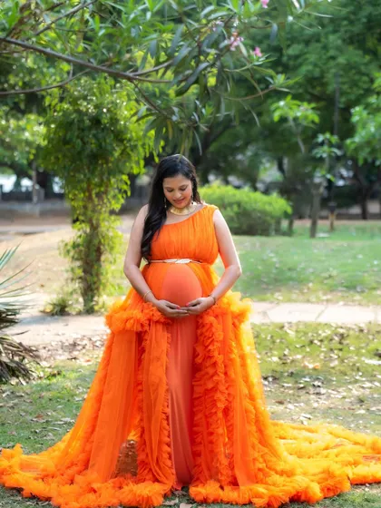A peaceful portrait of a mom-to-be in a vibrant orange gown, standing in a park and lovingly looking down at her baby bump.