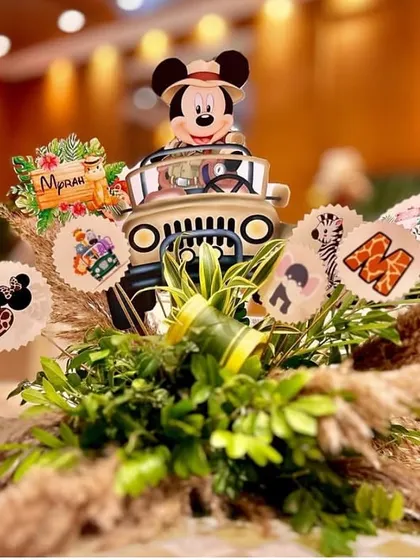 A creative jungle-themed table centerpiece. A miniature safari jeep with Mickey Mouse at the wheel is surrounded by lush greenery, pampas grass, and themed cutouts.