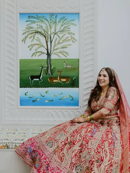 A happy bride in her red lehenga sits beside a colorful wall painting, her joyful laugh captured in a candid, relaxed moment.