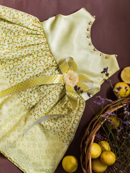 A fresh and sunny yellow dress featuring a delicate floral print and intricate eyelet lace panels. The waist is accented with a checkered yellow and white ribbon, adding a playful touch.