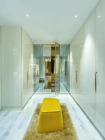 A spacious walk-in closet with sleek, handleless wardrobes and a central yellow ottoman on a plush rug. The glass-doored section allows for curated display.