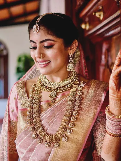 A smiling bride in her pink Kanchipuram saree, showing a close-up of her custom embroidered blouse and traditional temple jewelry.