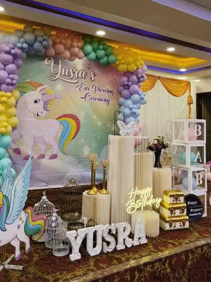 A magical unicorn-themed ear piercing ceremony and birthday party. This vibrant setup includes a large unicorn backdrop, pastel rainbow balloon arch, character cutouts, and a 'Happy Birthday' neon sign, creating a dream-like atmosphere.