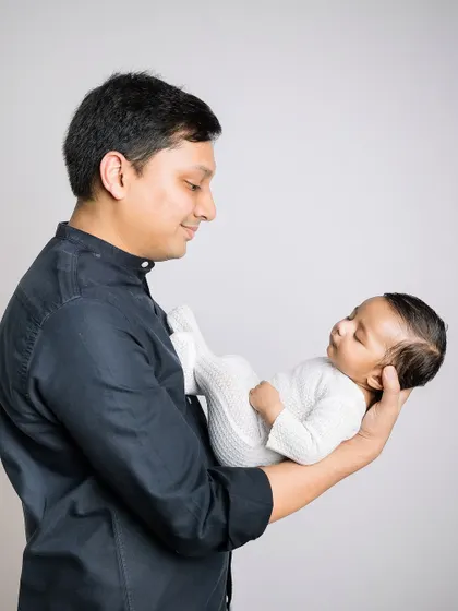 A father holds his sleeping newborn in one arm, looking down with a gentle, loving expression.