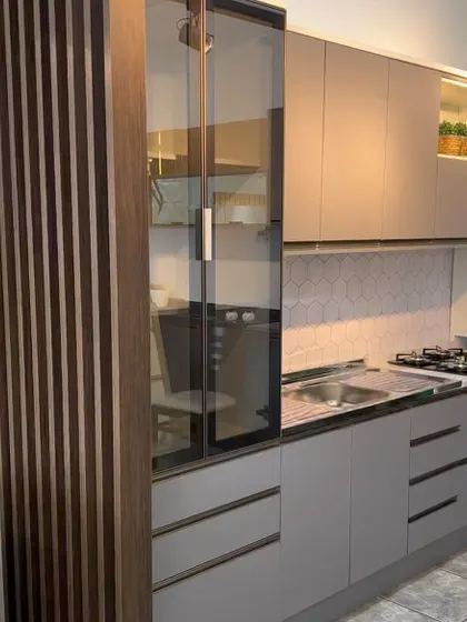 A modern kitchen design featuring matte grey cabinets and a tall glass-fronted display unit. The fluted wood partition on the side adds a textural element to the design.
