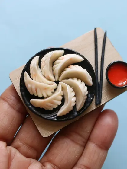 A handmade miniature of momos on a wooden board, complete with chopsticks and dipping sauce.