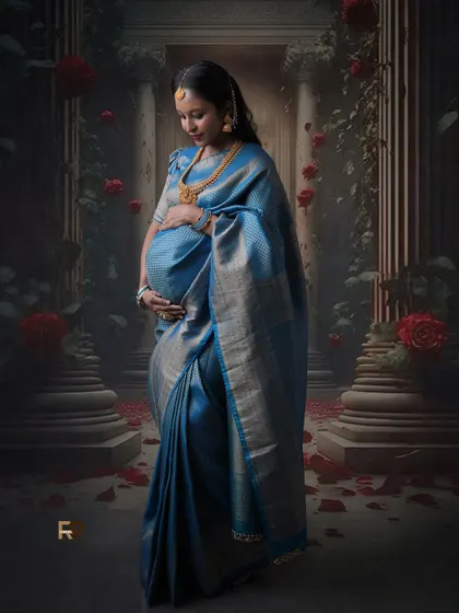 A majestic portrait. The mom-to-be looks regal in her blue silk saree, standing against a backdrop of grand pillars and roses, creating a truly royal feel.