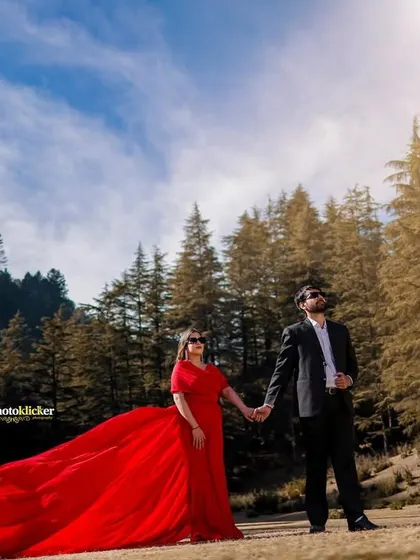 A dramatic, wide-angle shot of a woman in a flowing red gown holding hands with her partner in a sunlit forest clearing.