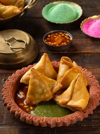 A Holi-themed shot of samosas served in a terracotta dish with chutneys. The background styling with colored powders adds a festive, celebratory context.