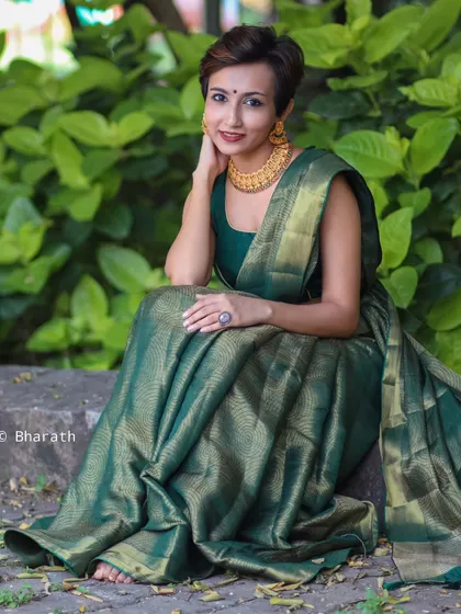 A full-length seated portrait that beautifully displays the drape and texture of the green silk saree in a natural, outdoor setting.