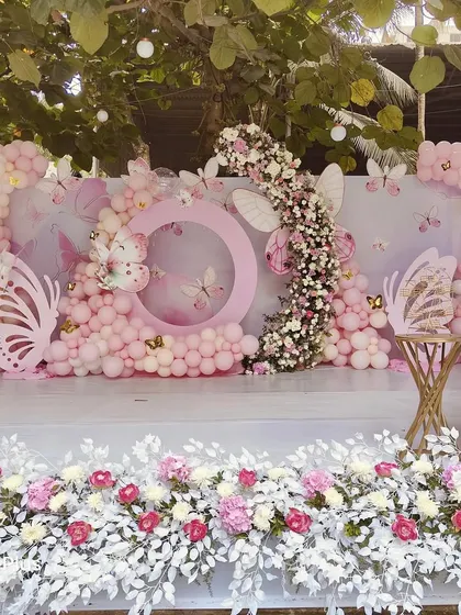A beautiful, wide shot of an outdoor butterfly-themed stage. The decor includes a full floral border at the front of the stage, a large balloon backdrop, and custom butterfly props.