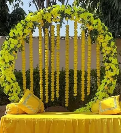 An outdoor Haldi seating area with a circular arch of yellow flowers and hanging marigold strings, creating a perfect photo spot.