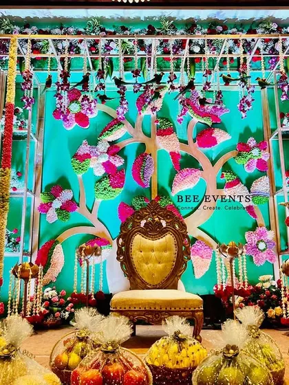 A vibrant and colorful stage setup for an engagement or pre-wedding ceremony. The backdrop features a 'tree of life' motif adorned with multi-colored flowers, surrounded by traditional elements and fruit baskets.