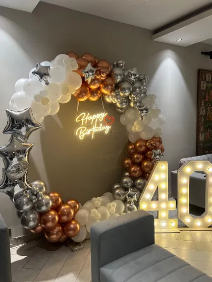 A 40th birthday decoration with a circular balloon frame in silver, white, and bronze. The setup includes a neon sign and large light-up marquee numbers.