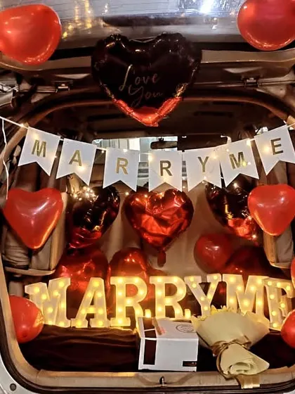 A creative and surprising car boot proposal setup. The trunk is decorated with a 'Marry Me' banner, heart-shaped balloons, and glowing marquee letters, creating an unforgettable and intimate moment for a marriage proposal.