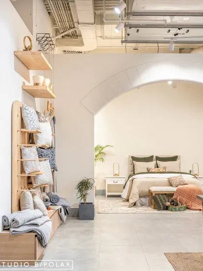 A lifestyle vignette of a fully dressed bed is framed by a graceful archway, creating an aspirational room-like setting within the store. The custom wooden shelving unit displays a variety of cushions and throws.