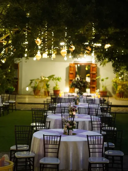 An enchanting garden dining setup. Tables are arranged under a canopy of trees adorned with hanging lanterns and fairy lights, creating a magical and intimate atmosphere for an outdoor reception.