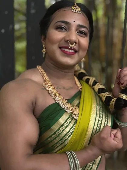 A smiling, close up shot in a green saree, holding my braided hair. This image combines traditional beauty with the visible results of dedicated strength training.