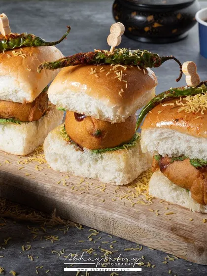 This image of Vada Pav captures a beloved street food in a clean, professional style. The styling with fried green chillis and chutneys adds authenticity and flavor cues.