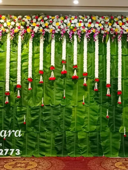 A traditional stage backdrop made entirely of fresh banana leaves. The look is completed with hanging garlands of white jasmine and colorful floral borders, perfect for baby showers and other ceremonies.