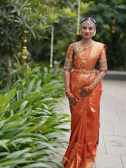 A bride in a vibrant orange and green Kanchipuram saree. The blouse features intricate embroidery that complements the saree's bold colors.