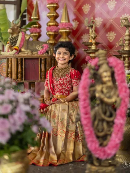 A joyful portrait of a young girl in her beautiful red and gold traditional dress, smiling brightly for the camera during her ear piercing ceremony.